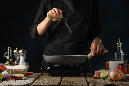 Chef in black uniform pours spices into the frying pan. Backstage of cooking traditional Indian chicken curry on dark blue background. Concept cooking process. Hot recipe. Frozen motion.