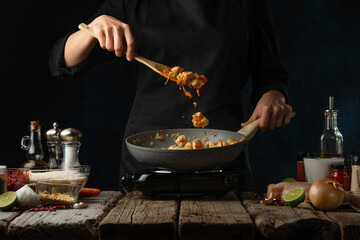 Chef in black uniform stir frying chicken fillet in the pan. Backstage of cooking traditional Indian chicken curry on dark blue background. Concept cooking process. Hot recipe. Frozen motion.