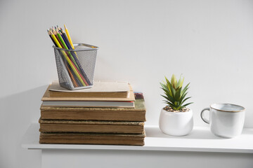Pencil holder with crayons on stack of old vintage books lying on mantelpiece. Artificial aloe in ceramic pot and porcelain cup