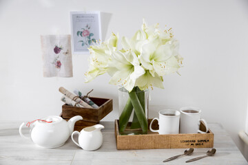 A bouquet of white lily in a glass vase on a table with two tall cups of coffee, a teapot, spoons, and a milk jug. Copy space