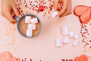 Beautiful young female hands put marshmallows in a cup of coffee, against the background of many small and large hearts . Valentine's Day concept. February 14, 2021 coming soon
