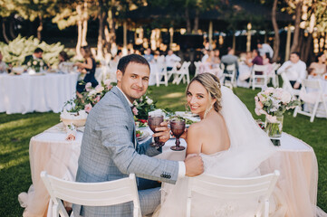 Stylish groom in gray plaid suit and blonde bride sit with faceted glasses of champagne at the...
