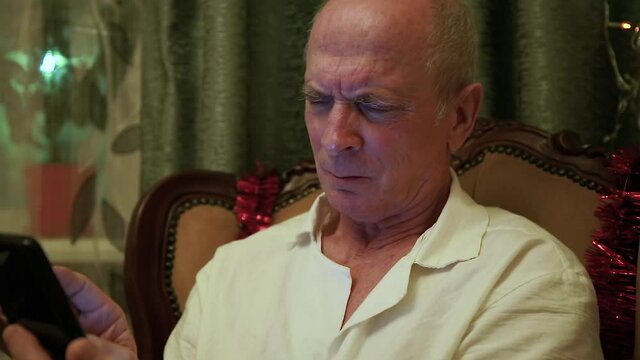 Happy elderly mac user 65-70 years old holding smartphone looking at screen, watching mobile video call online, relaxing at home, grandpa learning to use modern gadget technology.