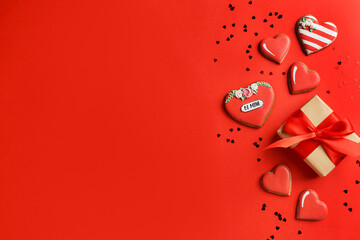 Cardboard gift box tied with a red satin ribbon with heart shaped gingerbread cookies and heart shaped confetti on red background. Valentine's Day or birthday greeting card. Top view, copy space