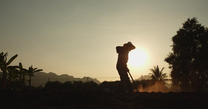 Slow motion silhouette scene of rural male Asian farmer digging the soil with a hoe to prepare for planting as the sun sets with sunlight passing through him beautifully.