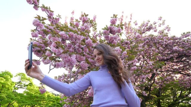European woman move long hair by hand and take selfie, she stand against flowering Japanese cherry tree at spring time. Tourist lady enjoy beautiful spring time at Tokyo city