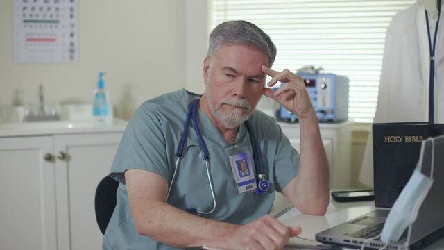 An overworked and discouraged doctor sitting at his desk in a small clinic picks up a Bible and starts reading it for encouragement and solace.