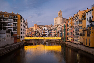 Fototapeta na wymiar Girona's urban cityscape skyline at dusk with famous gothic cathedral landmark and river houses reflected on a quiet river from red iron bridge