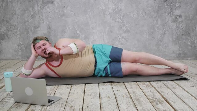 funny fat man doing sports on the yoga mat, doing sports online, looking into the laptop