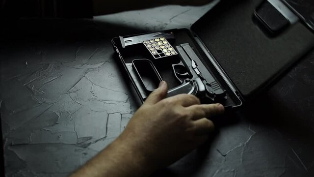 Person's hands open black briefcase with firearms and ammunition, insert clip with bullets in pistol and distort bolt. Preparing 9-mm gun for shooting on texture table.