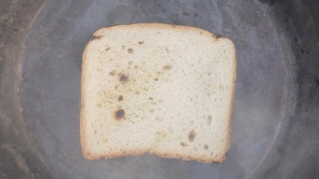 Timelapse overhead medium shot of a piece of sliced bread, cooking and burning inside an old pan, until it's completely destroyed and reduced to black char.
