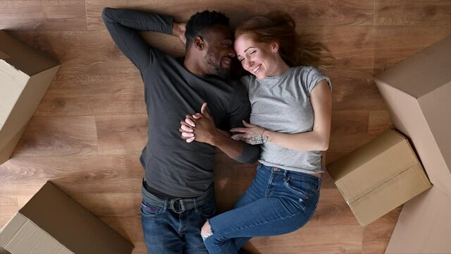 A interracial lovely couple lays on the floor in embrace among cardboard boxes and look to each other. A young family moved in a new house, apartment