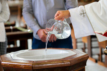 Priest pouring holy water into the baptismal font, moments before a child receives the sacrament of...