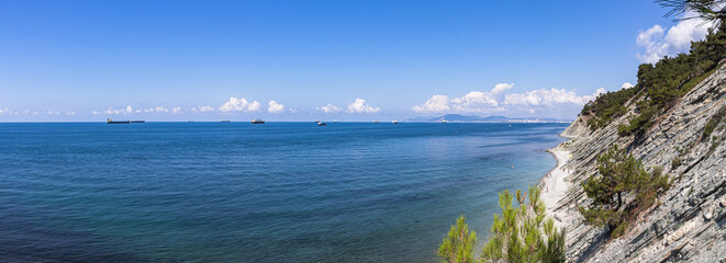 Panorama of the sea coast and wild beach at the foot of the rocks in the vicinity of the resort of Gelendzhik. Summer landscape, bright blue sky with clouds, tourists and cargo ships. Russia