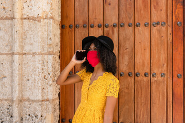 young black woman with curly hair, with a red mask, yellow dress and black hat, talking with her smart phone, in front of an old door