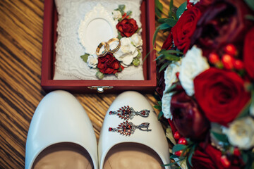 Vintage wedding jewelry and accessories, close-up. Gold wedding rings, ruby earrings, classic bride's shoes, bouquet of fresh red and white roses.