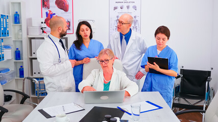 Mature doctor specialist briefing her medical team in conference room using laptop. Medical team, teamwork people discussing diagnosis about patients treatment issues record in workplace.