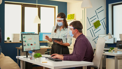 Business people wearing protective mask in new normal office making financial strategy pointing on desktop and taking notes on tablet. Multiethnic team working in company respecting social distance.