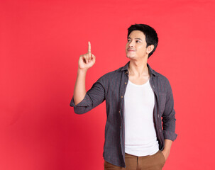 Young smiling attractive asian man in casual clothes posing isolated on red wall background, studio portrait. People sincere emotions lifestyle concept. Mock up copy space. Pointing index finger up