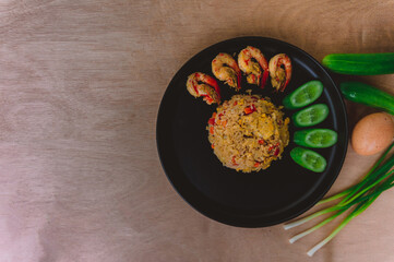 Shrimp fried rice in a black plate with spring onion, egg and cucumber all on the wooden table. top view.