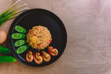 Shrimp fried rice in a black plate with spring onion, egg and cucumber all on the wooden table. top view.