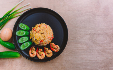 Shrimp fried rice in a black plate with spring onion, egg and cucumber all on the wooden table. top view.