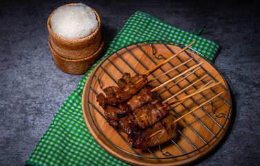 Grilled pork on bamboo skewers placed on a steel grill in a wooden tray with sticky rice in the cart, ready to serve