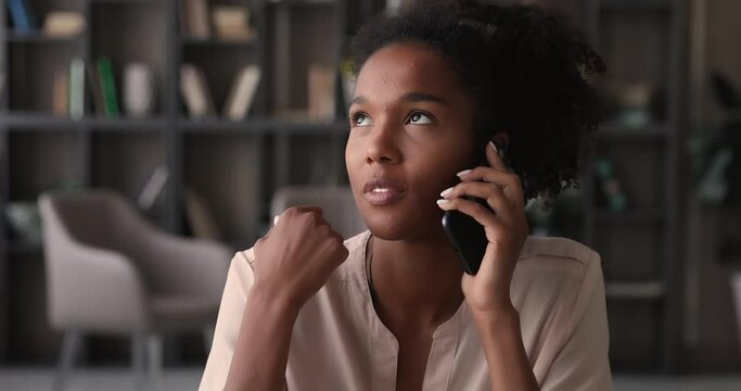 Attractive young african woman talk on phone sit at workplace enjoy formal or personal conversation. Mixed-race female holding modern smartphone solve business remotely, having pleasant blab concept