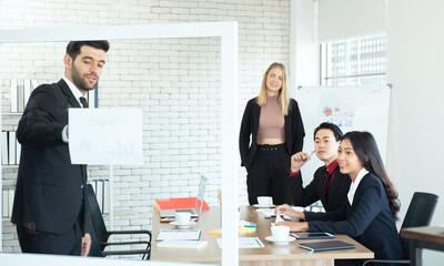 Asian business man and coworker woman look at caucasian manager giving presentation at work place.