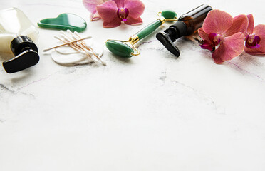 Face massage jade roller with cosmetic product on white marble background