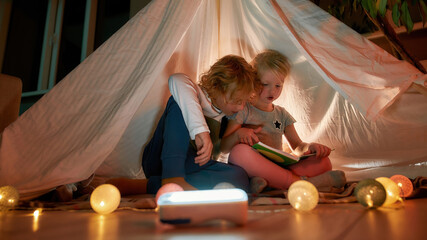 Obraz na płótnie Canvas Two little children, brother and sister reading a book together while sitting on a blanket in a teepee made with bedsheets at home