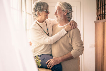 A moment of tenderness between two senior people who hug each other with love in front of the window, looking into each other's eyes. Concept of love and happiness