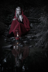 Fototapeta na wymiar Attractive woman dressed a little red riding-hood cross stream in a dark forest with a lantern