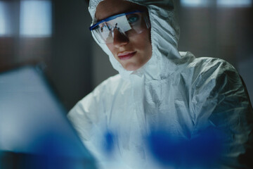 Female lab worker in protective overalls and goggles uses laptop at laboratory to analyze data. Viral vaccine researchers all over the world strive to create efficient anti-coronavirus treatment