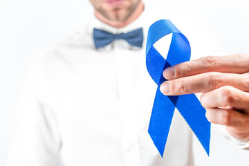 Blue ribbon cancer. Awareness prostate of health in November. Hipster men in bright shirt, cyan bowtie with blue ribbon in hands on white background. Symbol of oncology affected man. Copy space.