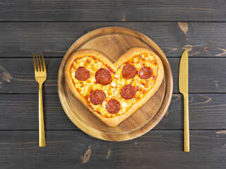 Pepperoni pizza with fork and knife on black wooden table. Flat lay. Top view. Love Italian food concept. Ready to eat heart shaped pizza. Black background.