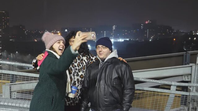 Small group of people two women and man adult family or friends on the roof in Manchester on New Year's night taking selfie photo while making funny faces. It is snowing and fireworks seen in distance