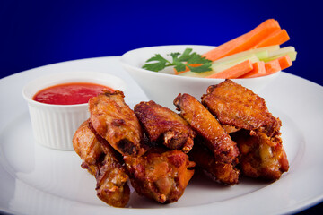 Roasted chicken wings with sticks carrots and celery and dipping sauce on white plate on blue background