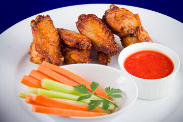 Roasted chicken wings with sticks carrots and celery and dipping sauce on white plate on blue background