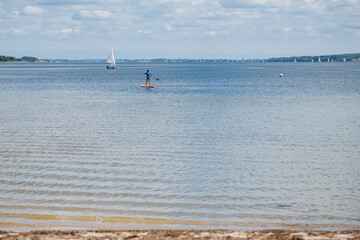 Woman paddling while standing. Blue sea with a sailboat