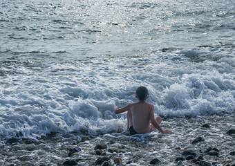 boy sits on the seashore. a boy in a swimming mask wants to swim underwater, but is afraid because of the strong waves in the sea. will he decide? boiling water, sun glare, wet stones on the shore