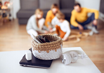 Mobile phones, laptops and digital tablets in a basket on table at home with playing family at back.