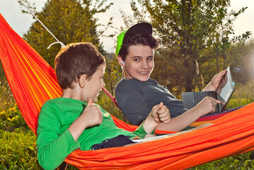 Two boys in orange hammocks on a green background. Children play with laptop and tablet. The concept of online learning in nature.