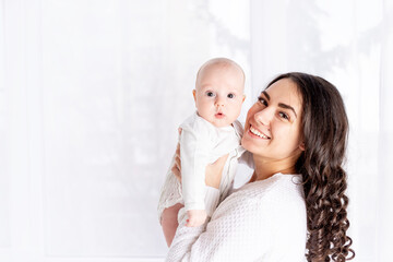 happy mother holding baby daughter at home by the window, mother's day, happy family concept, space...