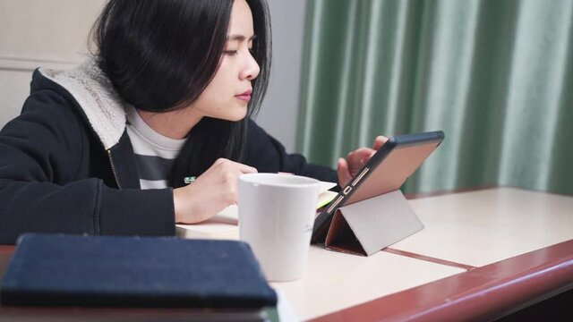 young asian female student, using tablet as educational media during lockdown, study at home, hand writing with pencil on notebook, wear sweater, distanced leaner, home school, online course lessons