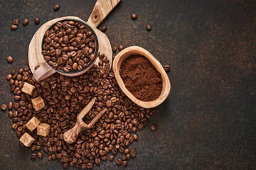 Coffee roasted beans in cup and scattered nearby, ground coffee and cane sugar on a brown table background. Top view with space to copy text.