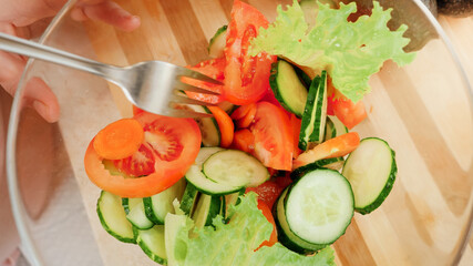Top view of woman mixing fresh vegetables and making tasty salad in big glass bowl on kitchen