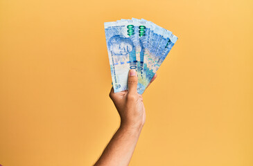 Hand of hispanic man holding south africa rand banknotes over isolated yellow background.