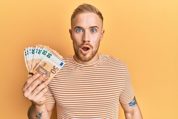 Young caucasian man holding bunch of 50 euro banknotes scared and amazed with open mouth for...