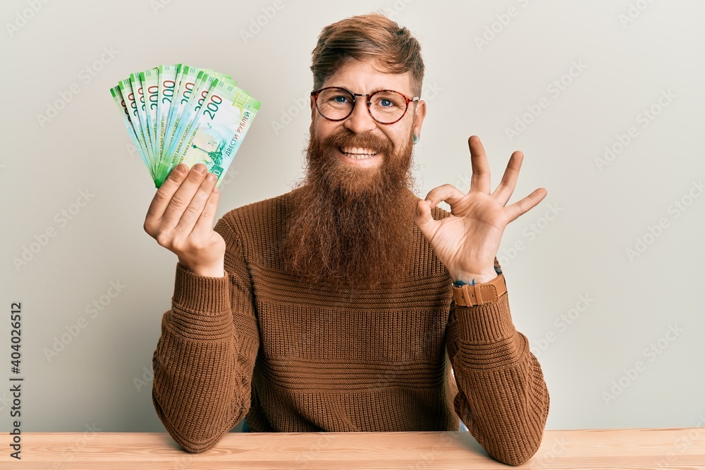 Sticker young irish redhead man holding russian 200 ruble banknotes sitting on the table doing ok sign with 